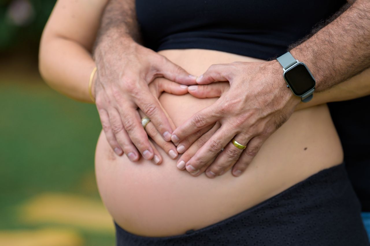 Close-up of a couple's hands forming a heart on a pregnant belly in an outdoor setting.